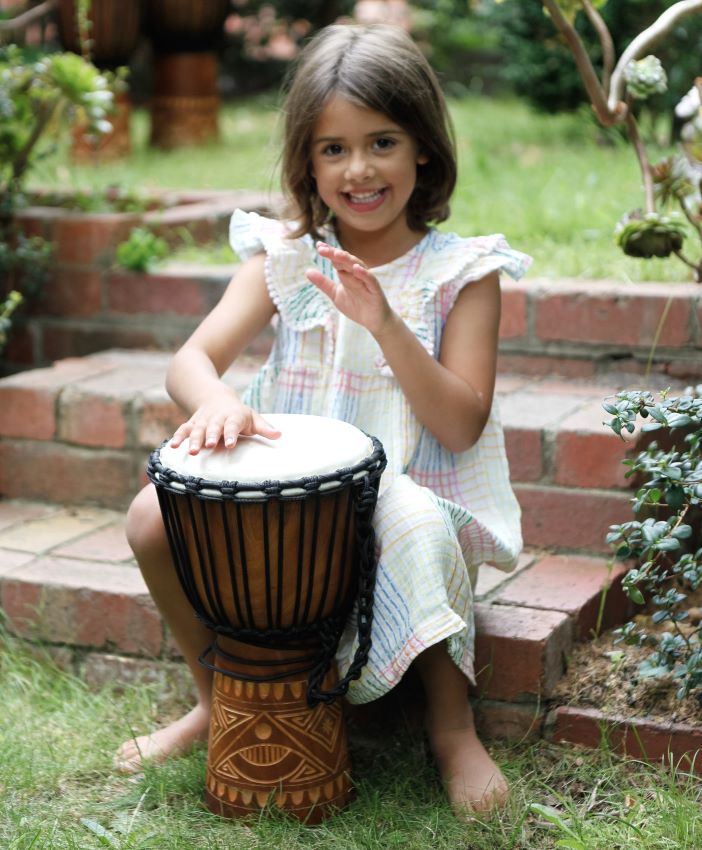 a chid playing a 30cm djembe while sitting in a backyard
