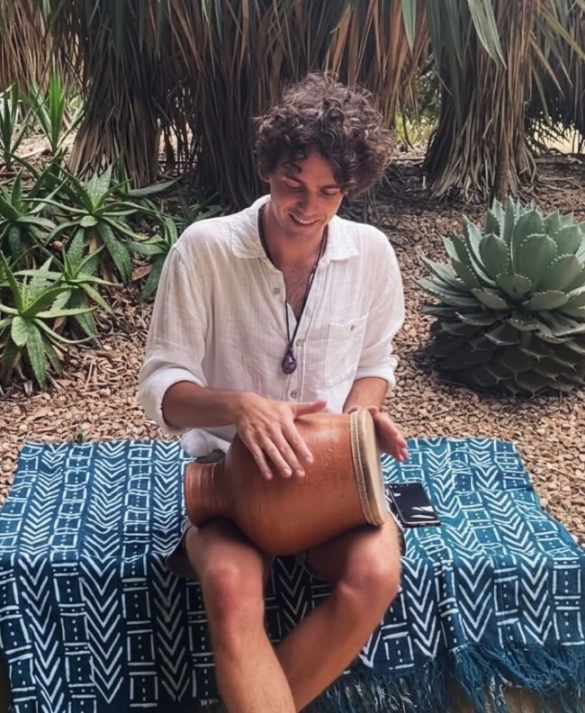 Person playing a clay udu drum seated outdoors among succulents and palms