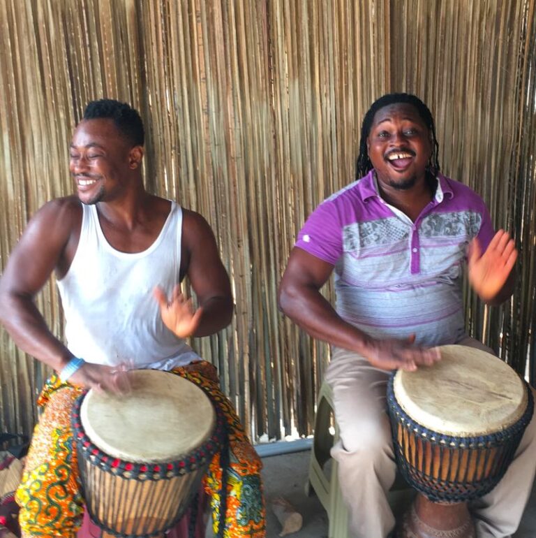 Two Ghanaian drummers playing djembes and sharing a joyful moment during a drumming session on the Rhythm Power Tour.
