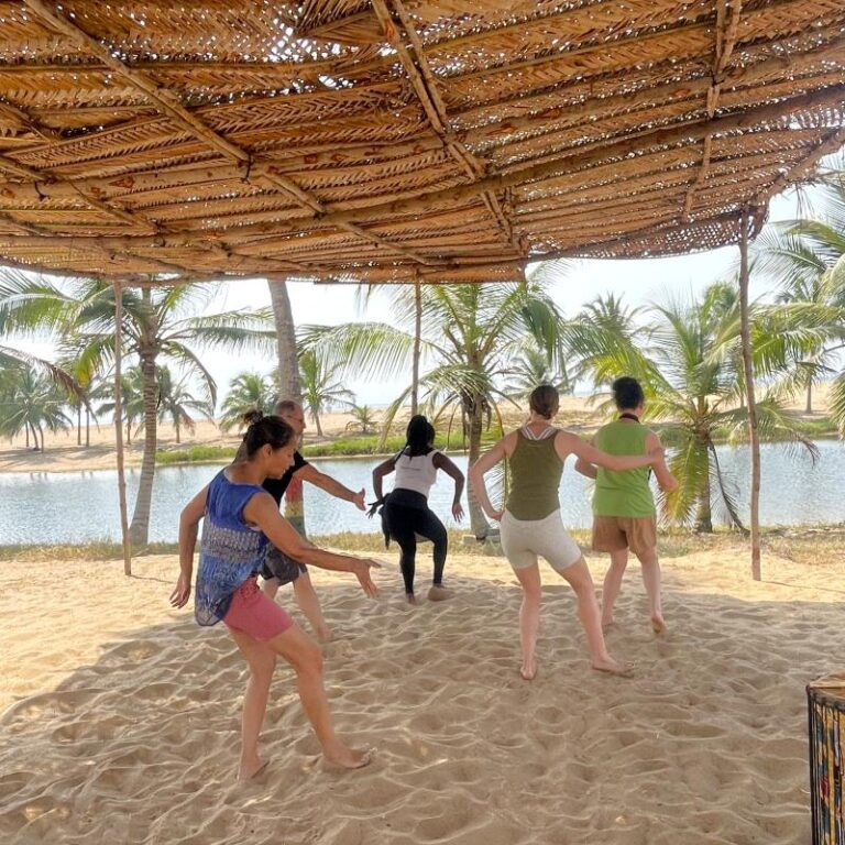 A group of participants dancing barefoot in the sand under a woven canopy during a West African dance class in Ghana.