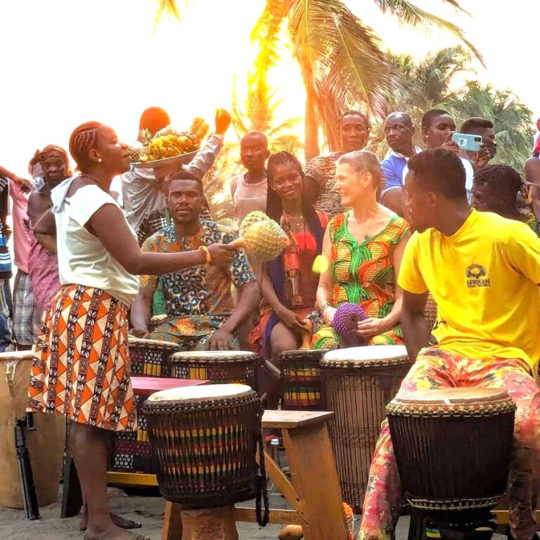 Community drumming gathering in Ghana at sunset, with local and international participants sharing music and shekere under palm trees.