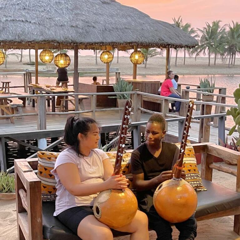 Kora lesson by the lagoon at sunset, with two people seated on a sandy deck in Ghana.