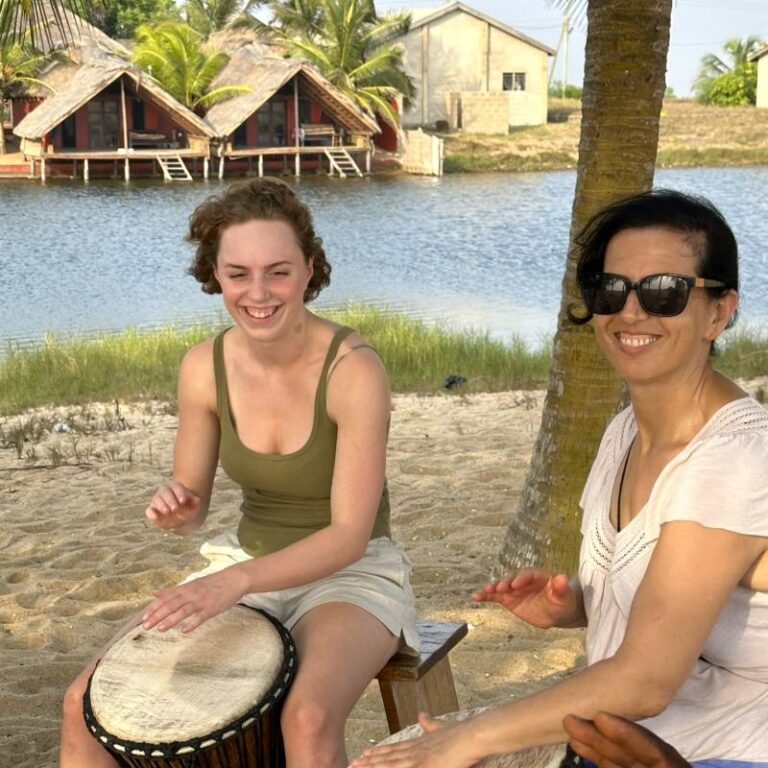 Two women drumming under palm trees on a sandy lagoon-side setting during the Rhythm Power Tour in Ghana.