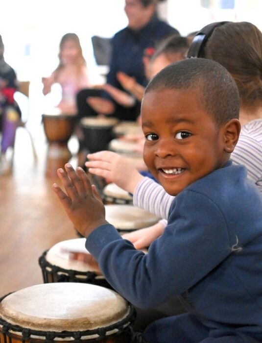 a preschool child smiling while playing a djembe in an african drumming incursion