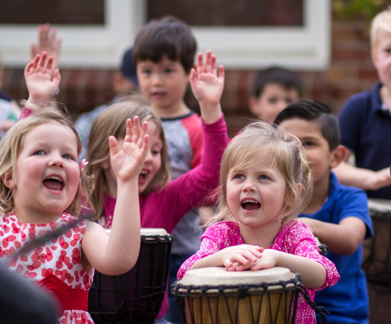 Children at a kindergarten African drumming incursion playing djembes, smiling and raising their hands during the music workshop.