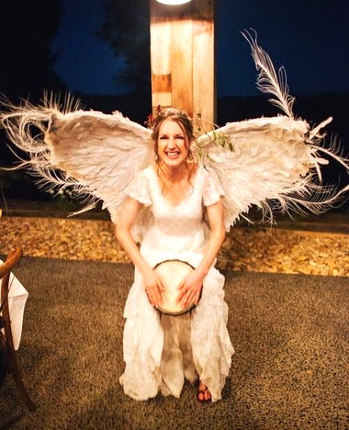 Bride in feather wings playing a djembe drum as live African drumming entertainment at a wedding