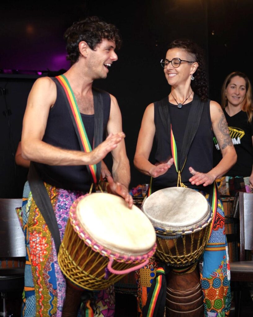 Drumming students performing on djembe drums at a Tribalism student concert in Melbourne, wearing colourful pants and smiling mid-play.