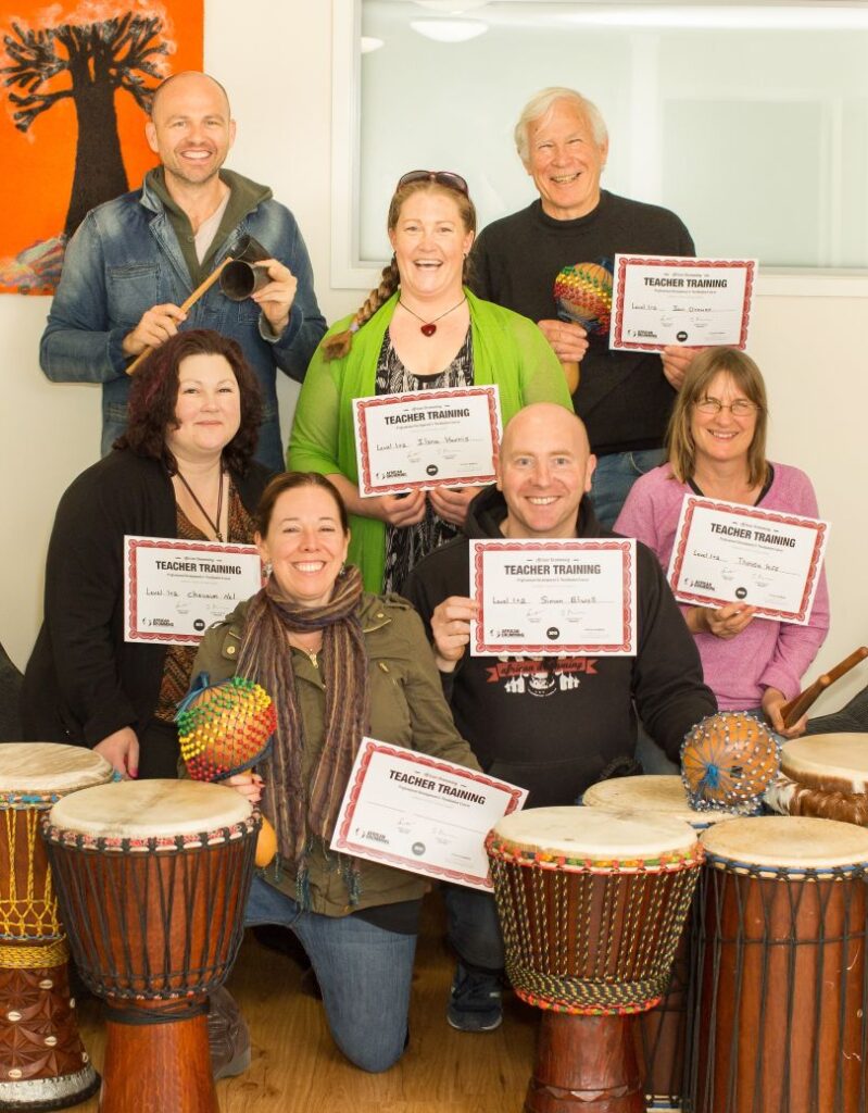 Group of facilitator training graduates holding certificates after completing African drumming training, surrounded by djembes and percussion instruments indoors.