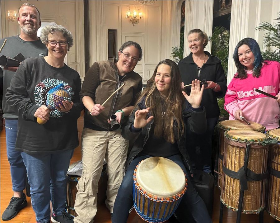 Group of participants at an African drumming facilitator training session, smiling while playing djembes, dununs, and percussion instruments indoors.