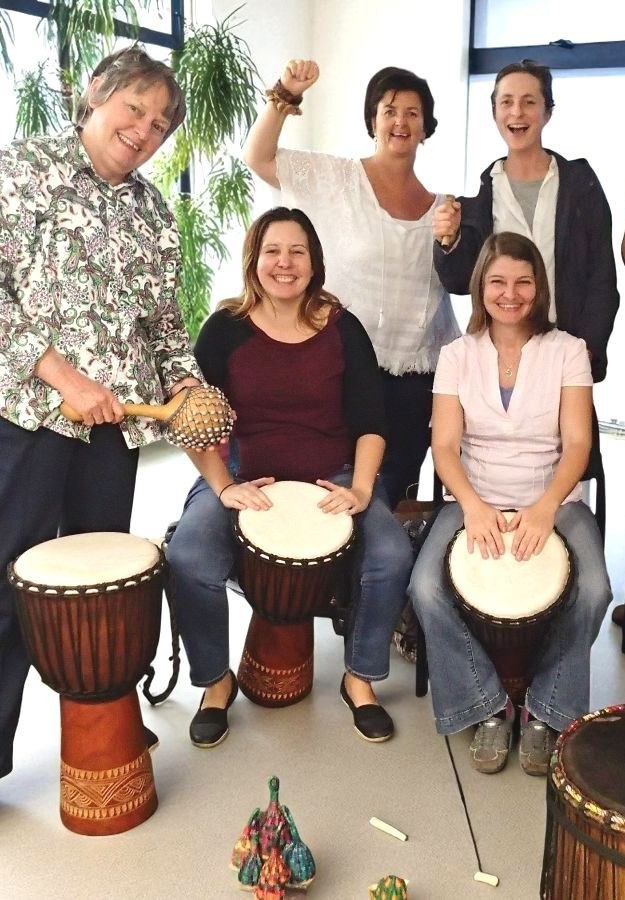Group of women learning African drumming during a facilitator training session, playing djembes and percussion instruments indoors.