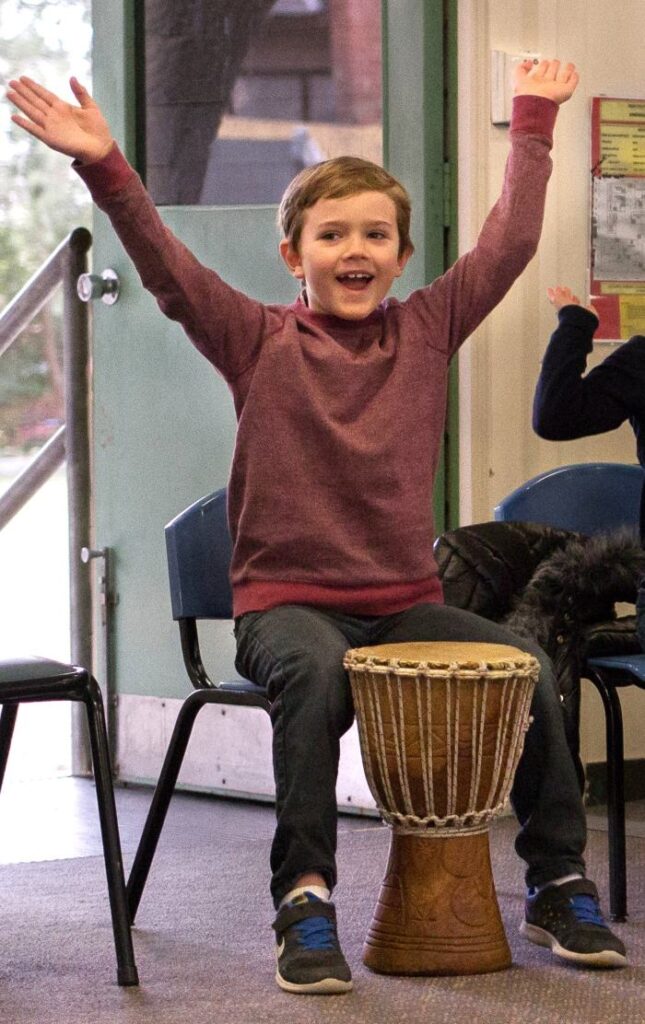 Primary school student smiling and playing a djembe drum during a classroom drumming session.