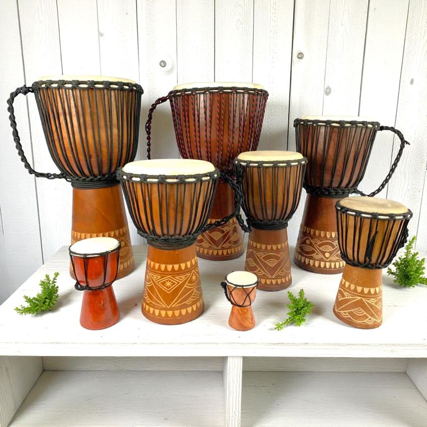 djembe drums in different sizes, from child to adult size, standing on a wooden shelf.