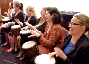 School staff sitting in a circle playing djembes, smiling and engaged during a team-building workshop.