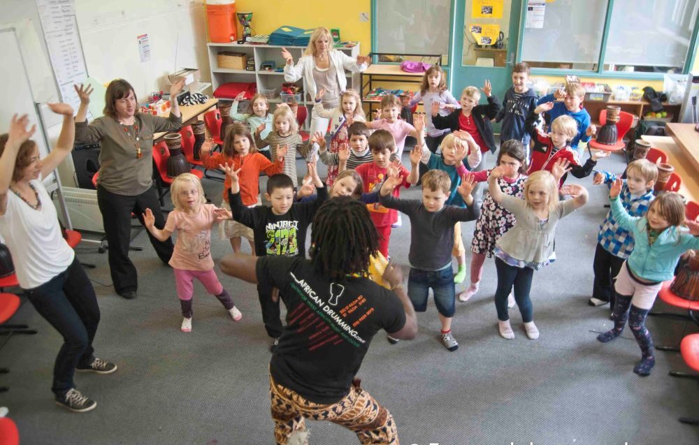 Kindergarten students dancing with their instructor during a West African dance workshop, raising their hands and moving together in the classroom.