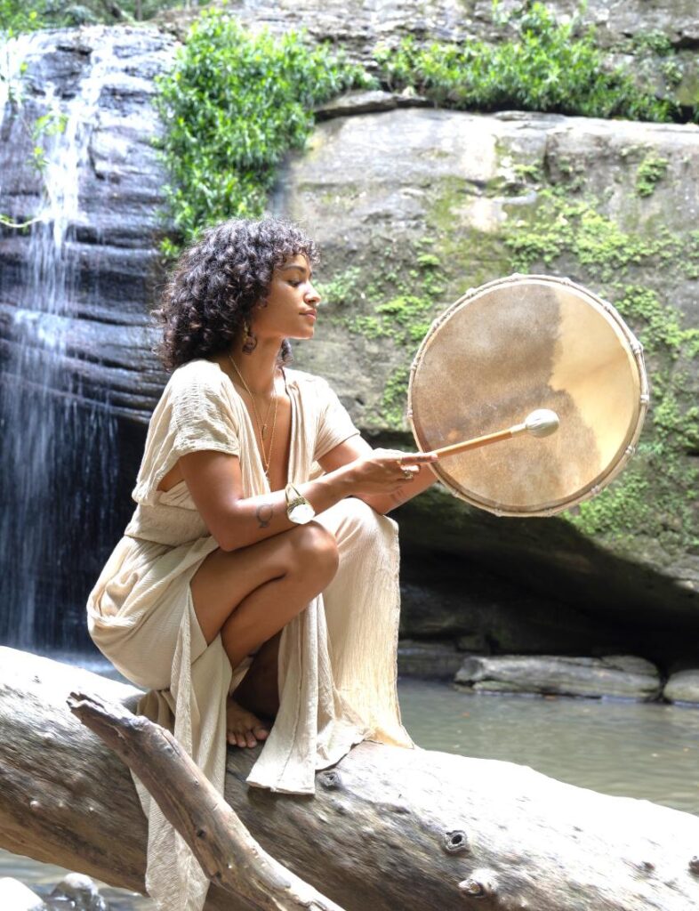 Woman playing a goat skin shamanic frame drum with a soft beater, seated on a log beside a waterfall