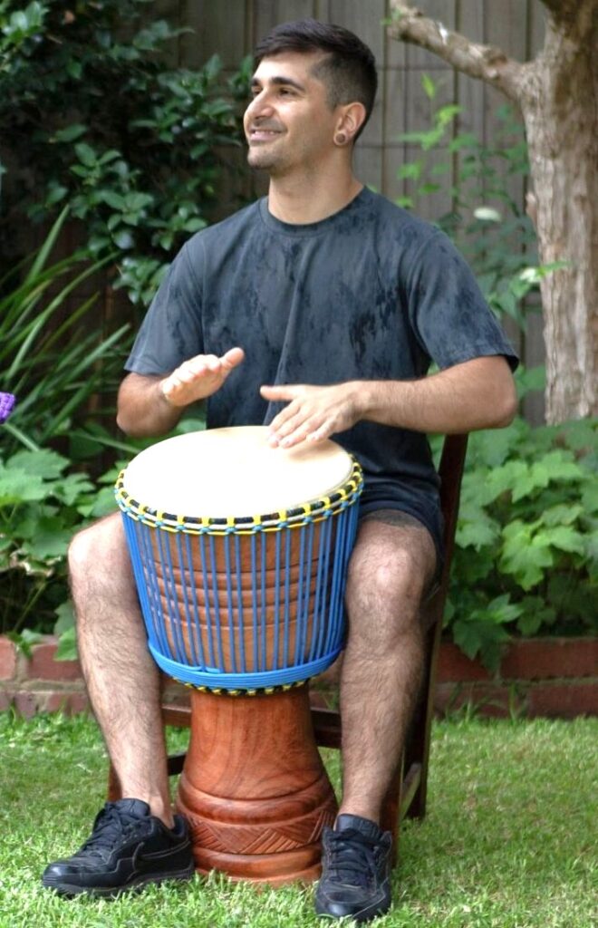Player seated with a Ghana djembe hand-carved from Twenaboa timber, with blue rope tuning and a natural skin head