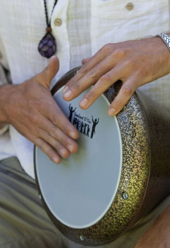 Hands playing a Gawharet El Fan darbuka with a speckled bronze finish, held sideways across the lap
